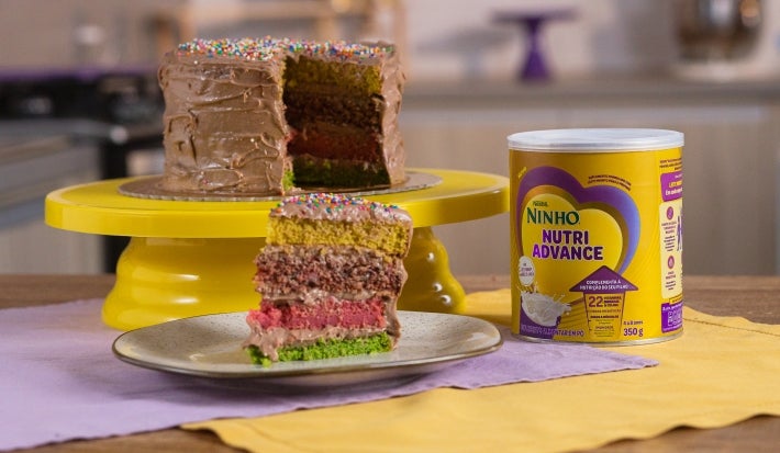 Fotografia em tons claros de um bolo inteiro sobre um apoio de bolo amarelo sobre dois panos, roxo e amarelo, em uma mesa de madeira. Ao lado do bolo, que está aberto com camadas coloridas, tem uma lata de NINHO Nutri Advance e uma fatia do bolo.