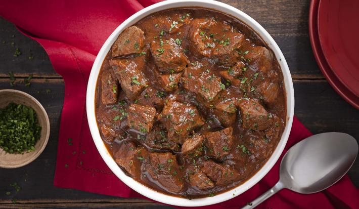 Fotografia em tons de marrom e vermelho, tendo ao centro uma panela com ensopado de carne e molho de cerveja preta, sobre guardanapo vermelho, ao lado um potinho com salsinha picada, uma colher de servir e uma tampa. Tudo sobre bancada de madeira rústica. Fotografia em tons de marrom e vermelho, tendo ao centro uma panela com ensopado de carne e molho de cerveja preta, sobre guardanapo vermelho, ao lado um potinho com salsinha picada, uma colher de servir e uma tampa. Tudo sobre bancada de madeira rústica.