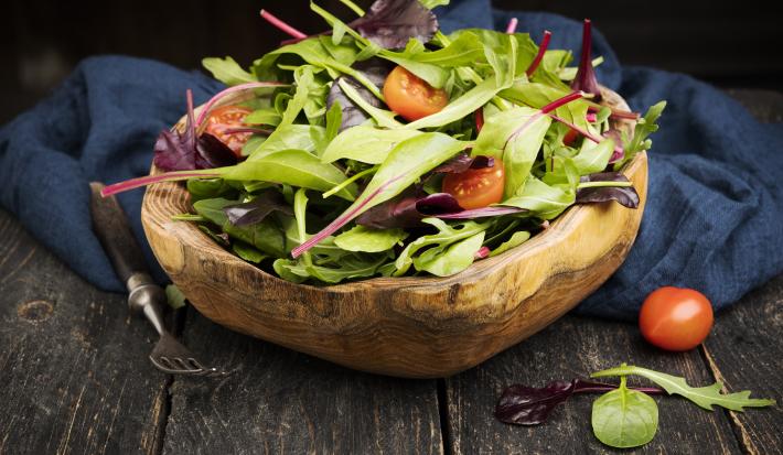 Fotografia em tons escuros de uma salada verde com salsão e tomates cereja dentro de um recipiente fundo de madeira, que está apoiado em um pano azul escuro, juntamente com um garfo de cabo preto, sobre uma mesa de madeira escura.