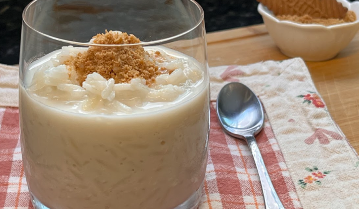 Imagem de um copo transparente ao centro com uma porção de arroz doce branco cremoso com uma farofa de biscoio triturado dourada por cima. o Copo está apoiado em cima de um pano xadrez vermelho e branco. Ao fundo, um biscoito Tostines.