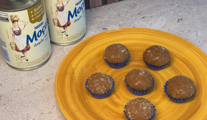 Seis brigadeiros caramelizados em forminhas azuis sobre prato amarelo, com latas de LEITE MOÇA® ao fundo.