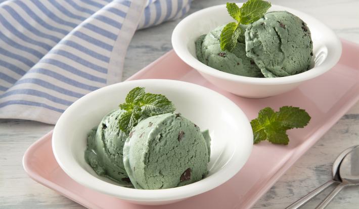 Foto da receita de Sorvete MOÇA de Menta com Chocolate. Observa-se dois potinhos brancos, cada um com duas bolas de sorvete decorados com folhinhas de hortelã, sobre uma tábua rosinha.