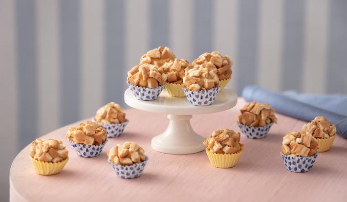 Fotografia em tons de rosa, branco e azul, tendo ao centro mini boleira com 5 docinhos envoltos em biscoito sobre toalha rosa, com outros docinhos espalhados ao redor, ao lado guardanapo azul, e fundo listrado em tons de azul.