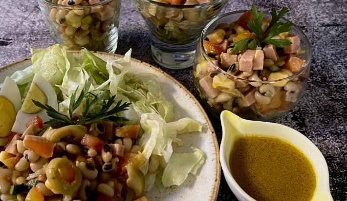 Foto da receita de Salada de Feijão Fradinho. Observa-se um prato grande com a salada e, ao lado, três potes individuais de vidro com a receita. Ao lado de tudo, um potinho com o molho de mostarda.