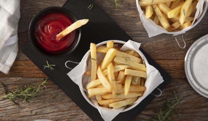 Fotografia em tons de marrom em uma bancada de madeira escura, uma tábua de ferro preta com um pote com batatas fritas dentro e um potinho ao lado com ketchup. Ao lado, um paninho e mais batata frita.