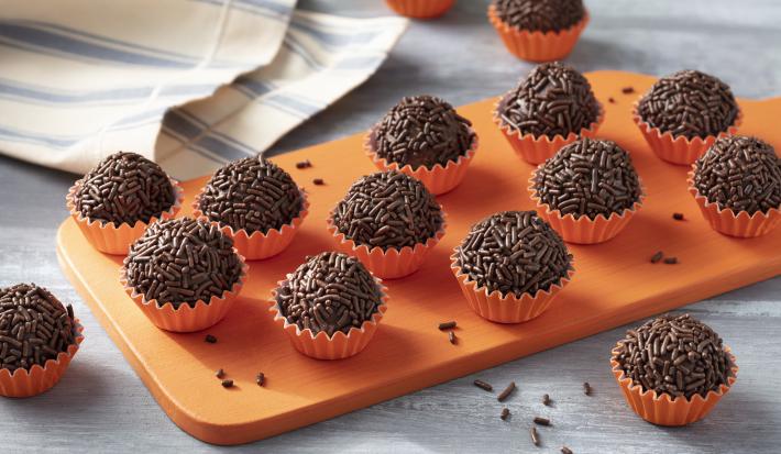 Imagem aproximada da receita de Brigadeiro Zero Lactose. Há diversos brigadeiros enrolados em forminhas em tom de laranja, todos sobre uma tábua de madeira em uma bancada decorada com um tecido em tom azul e bege Imagem aproximada da receita de Brigadeiro Zero Lactose. Há diversos brigadeiros enrolados em forminhas em tom de laranja, todos sobre uma tábua de madeira em uma bancada decorada com um tecido em tom azul e bege