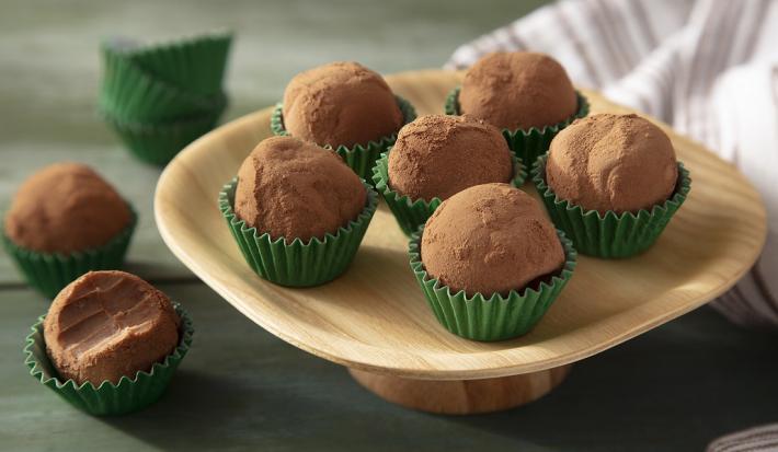 Fotografia em tons de marrom em uma bancada de madeira de cor verde. Ao centro, um prato de madeira contendo os brigadeiros. Ao, há um brigadeiro mordido e fundo, um pano branco.