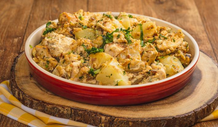 Foto de uma bancada de madeira. Ao centro há uma tábua de madeira sobre um pano quadriculado amarelo e branco e, sobre ela, um prato vermelho com a receita de frango e batatas gratinadas