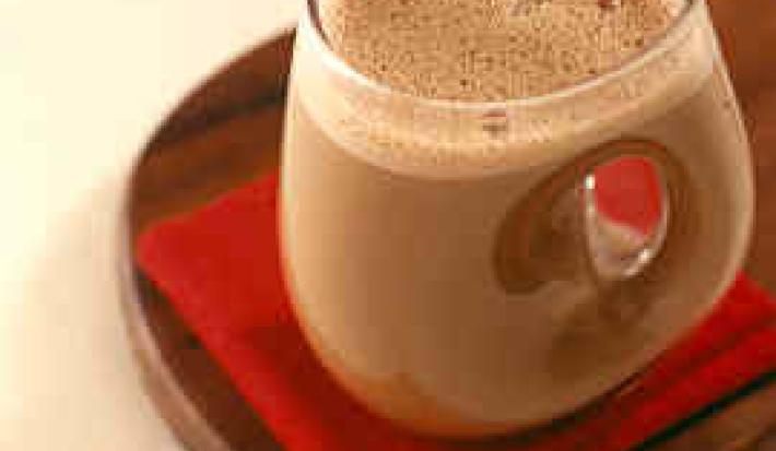 Fotografia em tons de vermelho em uma bancada de madeira, uma tábua de madeira escura, um paninho vermelho, uma xícara de vidro com a bebida quente feita com chocolate em pó.