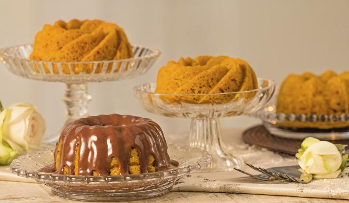 fotografia em tons de cinza, laranja e marrom vista de frente. Contém 4 bolos de cenoura e um deles esta com calda de chocolate, todos estão dentro de recipientes de vidros