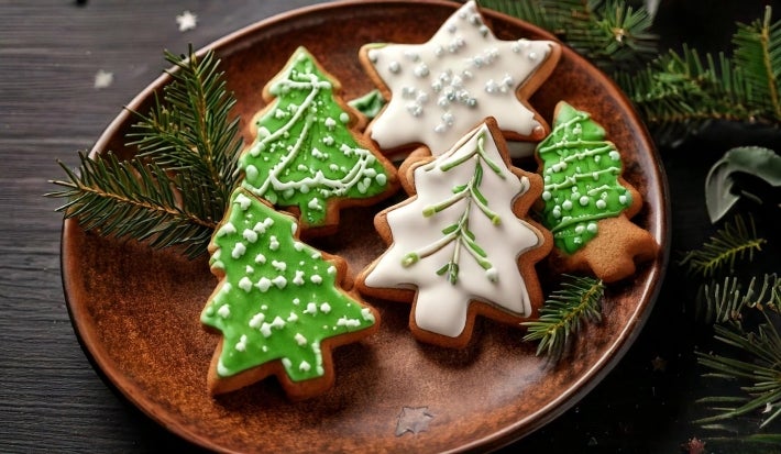 Em cima de uma mesa, prato redondo com biscoitos em forma de árvore de Natal e estrela, decorado com glacê branco e verde, e confeitos redondos brancos.