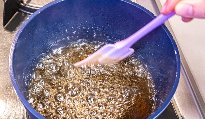 Foto aproximada da calda de caramelo fervendo em uma panela azul sendo mexida por uma espátula de silicone lilás