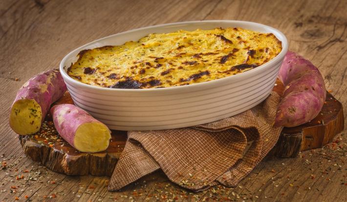 Fotografia em tons de marrom em uma bancada de madeira marrom. No centro, há um pano marrom com uma travessa branca em cima contendo o Escondidinho de Batata Doce com algumas batatas doces ao redor.