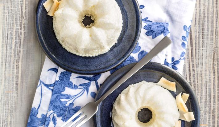 Fotografia em tons de azul em uma bancada de madeira clara, um pano branco com folhas azuis e dois pratinhos azuis escuros com o mini pudim de tapioca feito com iogurte, abacaxi e coco.