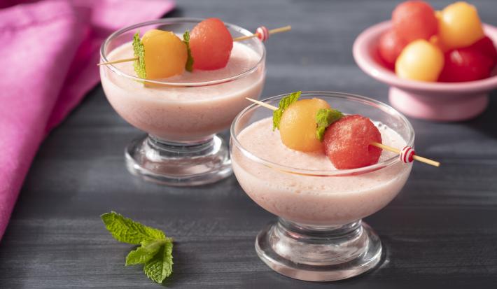 Fotografia em tons de rosa em uma bancada de madeira escura, um paninho rosa, duas taças de vidro com o creme de iogurte de morando com espetinhos de fruta em cima dele. Fotografia em tons de rosa em uma bancada de madeira escura, um paninho rosa, duas taças de vidro com o creme de iogurte de morando com espetinhos de fruta em cima dele.