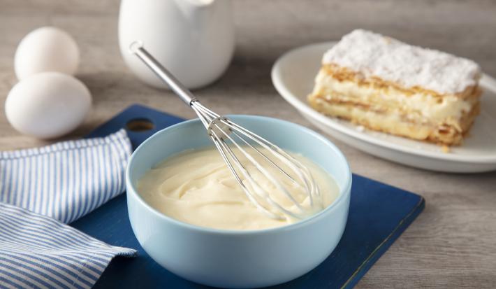 Fotografia em tons de azul em uma bancada de madeira, um pano azul listrado, uma tábua de madeira azul, um bowl azul claro com o creme belga dentro e um fouet. Ao lado, um prato com um pedaço de uma torta com o creme belga.