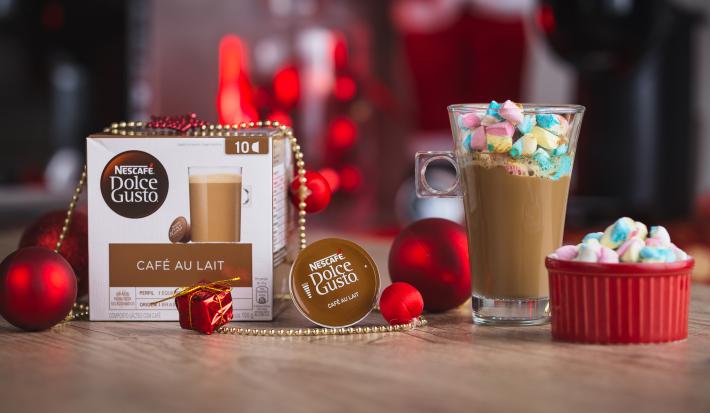 Fotografia em tons de vermelho em uma bancada de madeira, uma xícara de vidro alta com a bebida natalina dentro e um vidrinho vermelho com vários marshmallows coloridos. Ao fundo, decoração de natal.