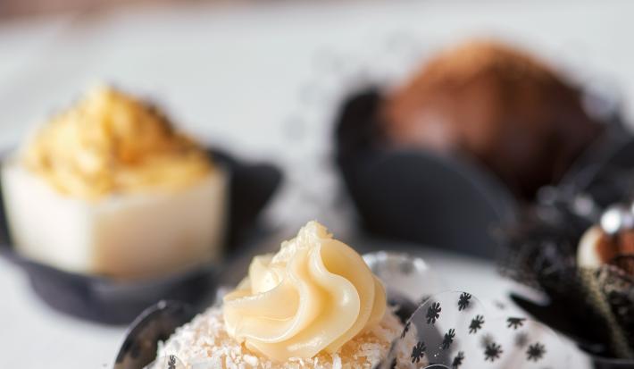 Fotografia de alguns docinhos de banana decorados. Ao fundo, docinhos de chocolate também decorados, sobre uma mesa de madeira em tom claro.