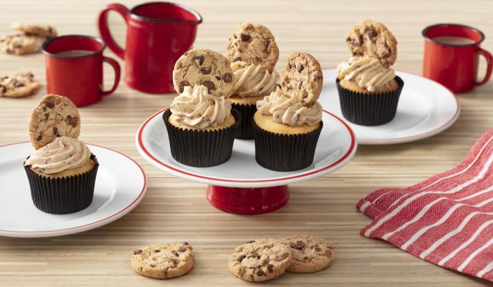 Foto de uma bancada clara com canecas e um bule vermelhos, um tecido listrado vermelho e branco. Há dois pratos brancos com cupcakes em cima e, ao centro, um prato com três cupcakes decorados com cookies.
