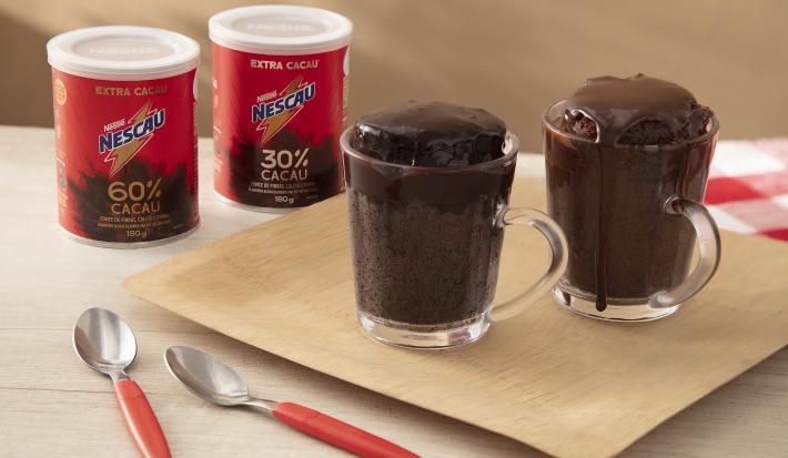 Foto da receita de Bolo de chocolate na caneca servida em duas canecas de vidro sobre uma tábua de madeira com duas latas de nescau extra cacau e duas colheres de decoração