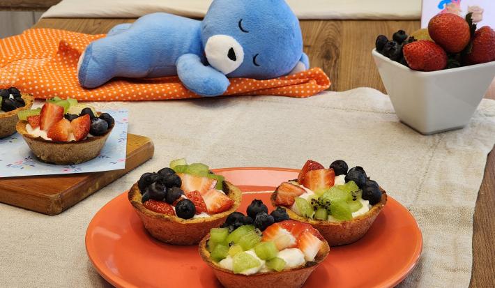 Fotografia de um prato laranja no centro da foto com três cestinhas de mucilon com frutas e iogurte. Ao lado tem uma mini tábua de madeira com mais duas cestinhas dessas. No fundo da foto tem um paninho laranja, o ursinho azul da marca e frutas.