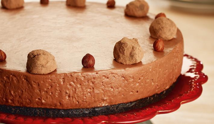 Fotografia em tons de chocolate, grafite e vermelho, tendo ao centro torta de chocolate sobre boleira vermelha e guardanapo grafite; ao fundo pratos empilhados com um talher por cima, tudo sobre bancada de madeira.