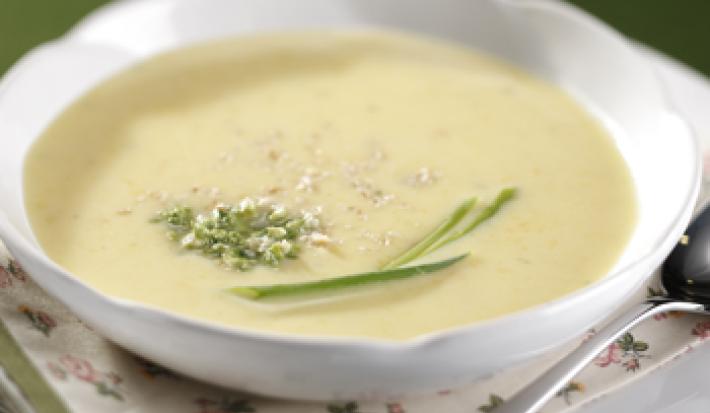 Fotografia em tons de verde em uma bancada com uma bandeja branca, um pano branco com detalhes verde e em flores, um prato redondo branco fundo em cima dela e o creme de mandioquinha.