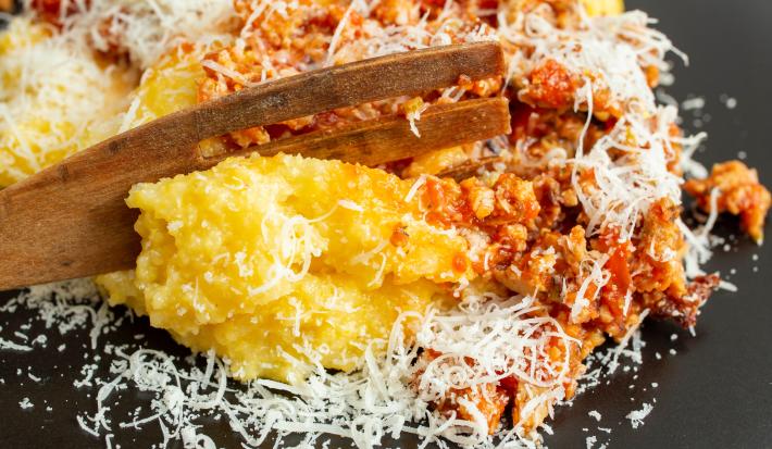 Fotografia de um prato raso preto com uma polenta gratinada recehada com carne moída e queijo ralado por cima. Apoiado na receita, tem um talher de madeira grande.