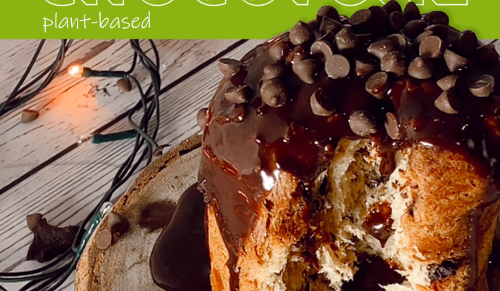 Foto em tons de marrom da receita de chocotone plant-based servida com calda de chocolate vegano sobre uma tábua de madeira redonda. Ao fundo, há uma embalagem de bebida vegetal de castanha de caju natures heart