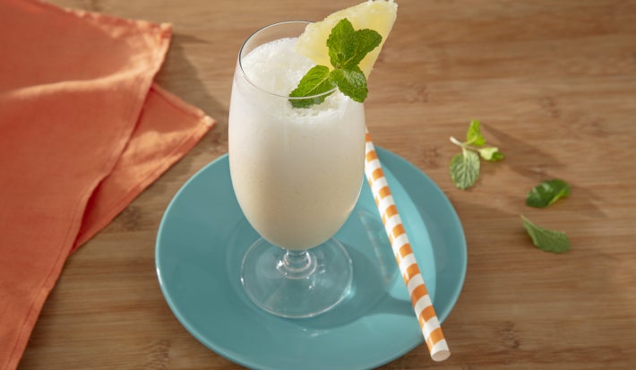 Foto da receita de pina colada feita com novasource proline servida em um copo de vidro para drinks com um canudo listrado ao lado sobre um prato azul claro