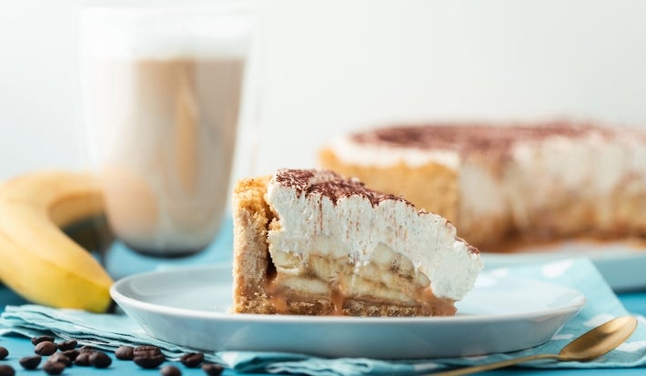 Em cima de uma mesa, fatia de banoffe com massa crocante, decorado com chantily e canela em pó.