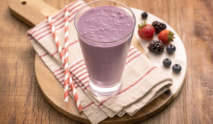 Foto em tons de roxo da receita de milkshake de uva com frutas vermelhas servida em um copo alto sobre uma tábua de madeira um pano listrado vermelho e branco com canudos de mesma cor e frutas vermelhas usadas na receita ao lado como decoração.