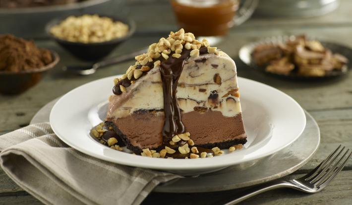 Fotografia de uma fatia de torta Alaska feita de sorvete de café com uma calda de chocolate e amendoins triturados. A torta está em um prato fundo branco.