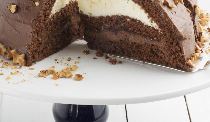 Fotografia em tons de marrom em uma bancada de madeira clara, um suporte de bolo branco com pé preto e uma doce de chocolate com recheio de creme branco e de chocolate.