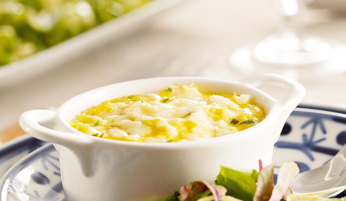 Fotografia em tons de verde e azul em uma bancada de madeira branca, um prato branco com detalhes azuis ao centro e salada verde em cima dele, com um potinho branco e a brandade de bacalhau dentro dele.