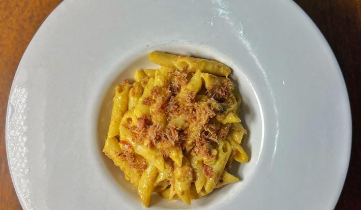 Fotografia vista de cima de um prato de vidro branco com bordas grossas e fundo pequeno sobre uma mesa de madeira. Dentro do prato tem uma receita de penne cremoso com farofa de bacon.