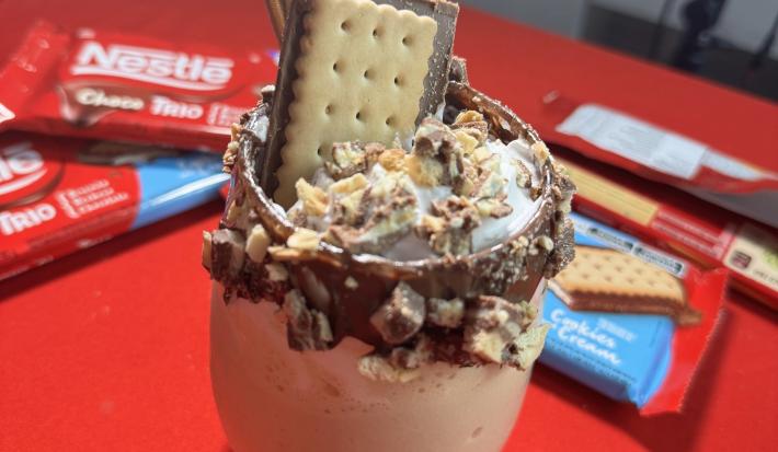 Fotografia de um copo de vidro com a borda com creme de avelã, pedaços de chocotrio, chantilly e uma unidade inteira de chocotrio. Dentro do copo tem milkshake. O copo está sobre uma mesa vermelha, com quatro embalagens de chocotrio.