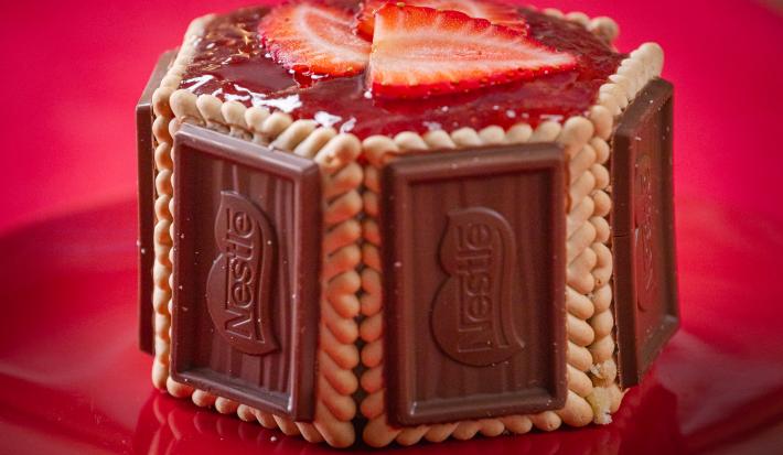 Fotografia em tons de vermelho com uma sobremesa ao centro. Essa sobremesa é feita por camadas laterias de biscoito de chocolate Choco Biscuit e finalizadas com geleia de morango no topo