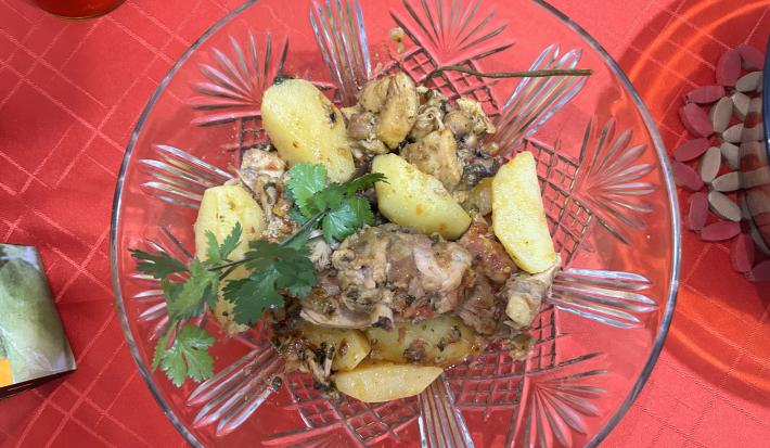 Fotografia vista de cima de um recipiente fundo de vidro com frango cozido, batatas e coentro por cima. O recipiente está sobre uma toalha de mesa vermelha.