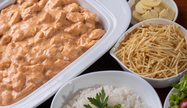 Fotografia de uam travessa branca retangular com o strogonoff de frango, ao redor tem três recipientes. Em cada um tem o champignon fatiado, batata palha e arroz branco.