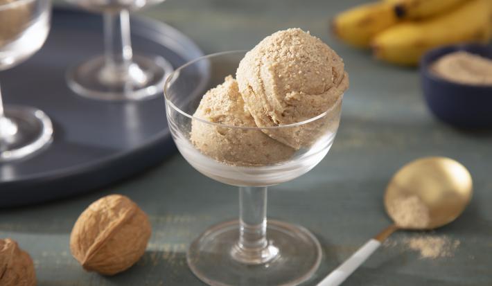 Foto da receita de Sorvete Crocante de Farinha Láctea Nestlé. Observa-se uma taça de sobremesa de vidro transparente com duas bolas do sorvete. A foto é decorada com Farinha Láctea, nozes com casca e bananas