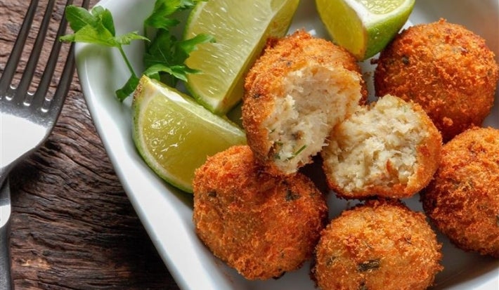 Em cima de uma mesa, recipiente branco com porção de bolinhos de bacalhau dourados e crocantes e lascas de limões. Centralizado, há um bolinho aberto ao meio com bacalhau cremoso.