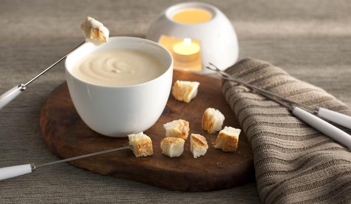 fotografia em tons de cinza, marrom e banco de uma bancada vista de frente. Contém uma tábua redonda de madeira com um recipiente redondo e branco que contém o fondue, 4 garfos para fondue e pedaços de pães ao redor
