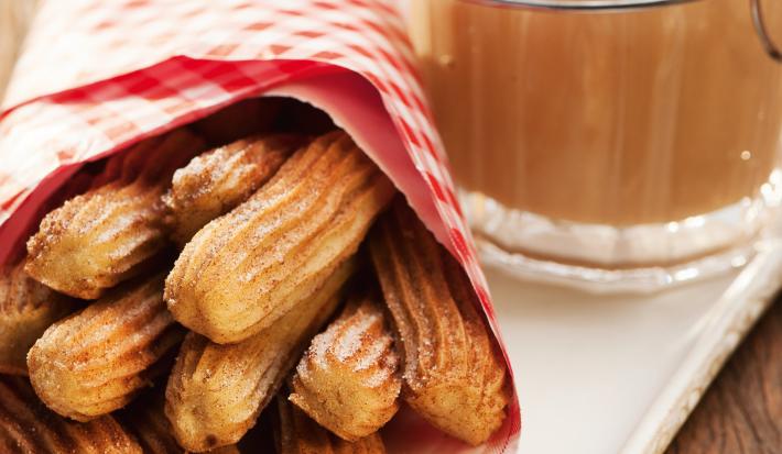 Fotografia em tons de marrom e branco de uma bancada de madeira com um prato retangular, sobre ele um paninho vermelho com churros. Ao lado um recipiente com doce de leite.