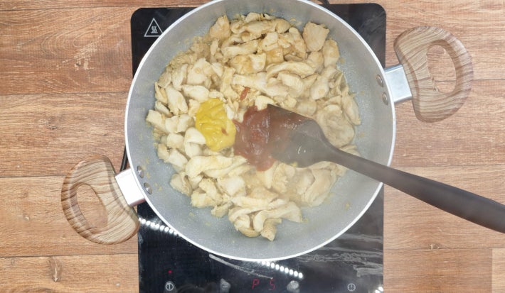 Frango com Ketchup e Mostarda dentro de uma panela com uma espátula de silicone, apoiado sobre fogão portátil preto.