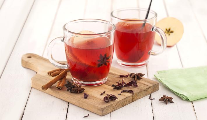 Fotografia em tons de verde em uma bancada de madeira branca, um paninho verde, uma tábua de madeira, duas xícaras de vidro com o chá de hibisco com maçã dentro deles. Ao lado, paus de canela, cravos e anis-estrelado.