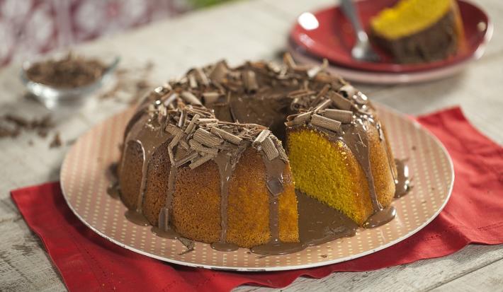 Imagem de um bolo de cenoura com cobertura de brigadeiro e raspas de chocolate sobre um prato rosa claro de bolinhas brancas. Ao fundo, um prato com uma fatia de bolo. u