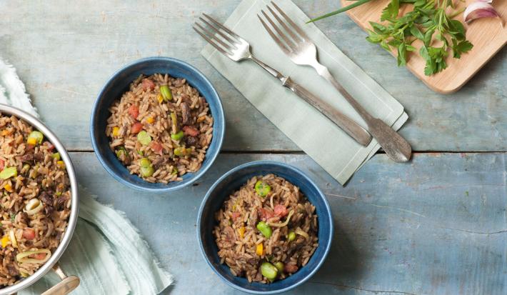 Fotografia em tons de azul em uma bancada de madeira azul com um pano branco com listras azuis, um panela com arroz e carne. Ao centro, dois potes redondos fundos com o arroz, carne e vegetais dentro dele.