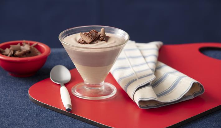 Foto de uma bancada sobre a qual há uma tábua vermelha com uma taça em que está a receita da mousse, dividia em uma camada de mousse branca e outra de mousse de Nescau. Há também um guardanapo de tecido, uma colher de sobremesa e um pote com raspas