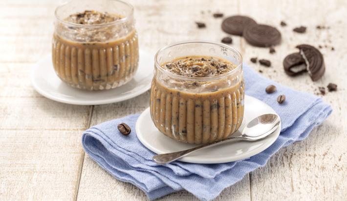 Foto da receita de Palha Italiana de Colher. Observa-se duas taças redondas e transparentes com a palha italiana de colher em um pires branco em cima de um guardanapo azul claro sobre uma superfície de madeira. Atrás, biscoitos NEGRESCO de decoração.